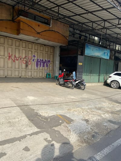 The image shows a street scene in Purwakarta, Indonesia, with a man sitting on a red scooter. The scooter is parked in front of a closed shop with large, beige, wooden, double doors with graffiti on them. The shop has a covered overhang. The man is facing the camera and wearing a red shirt, a dark cap, and a blue face mask. There are plastic chairs next to the scooter. A white car is parked on the right side of the image, further down the street. The street is paved with grey concrete with some cracks and patches of black asphalt. A chain is strung across the road, which appears to be a driveway or entrance to the shop. In the background, behind the shop, there are a green, wooden gate and a blue sign with white text. There is also a small, yellow, caution sign posted on the building. The overall scene appears to be in a commercial area of the city.
