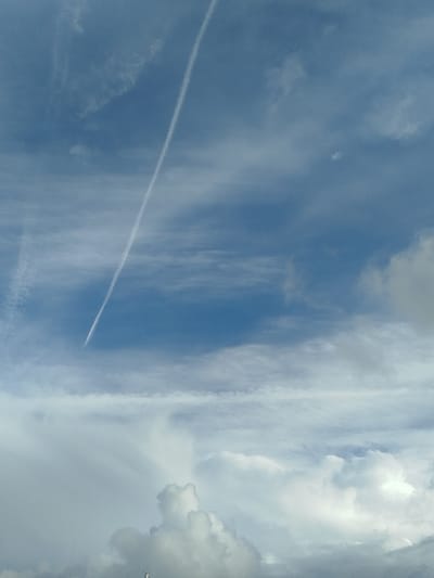 A vertical, white contrail from an aircraft cuts diagonally across a clear blue sky in the upper half of the image. Below the contrail, horizontal bands of wispy clouds stretch across the sky. In the bottom third of the image, dense, fluffy white clouds fill the frame, obscuring the ground. A small, dark object resembling a utility pole or antenna with a few horizontal protrusions is visible at the very bottom center of the image, suggesting it is part of the landscape below the clouds. The overall scene depicts a sky with varying cloud formations and a prominent airplane contrail. The location in the city of Ko Kaeo, Thailand, is not discernible from the visual information present in the image.