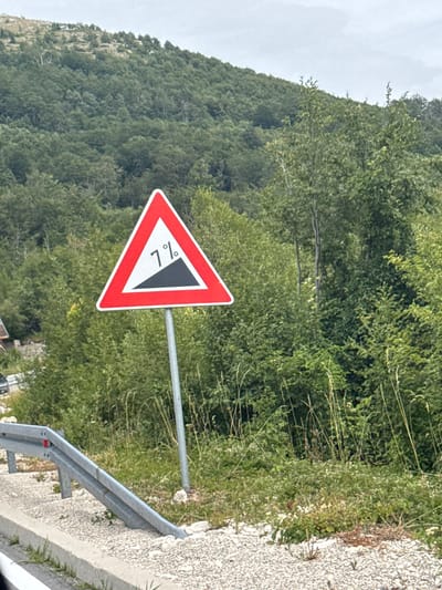 An upright, triangular traffic sign with a red border and white interior is posted on a metal pole. The sign depicts a black, upward-sloping ramp with the number "7%" in white text positioned to the left of the ramp. The sign is located beside a road in a rural, hilly area with dense green foliage and trees covering the background. A metal guardrail is visible in the foreground, angled downwards. The ground is covered in gravel and some sparse vegetation. The sky is overcast. There are no people visible in the image.