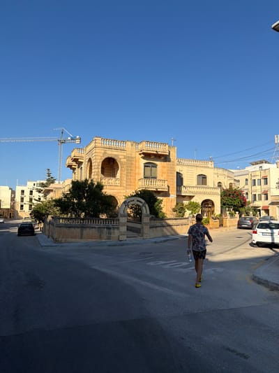 A man wearing a colorful short-sleeved shirt and patterned shorts walks away from the camera down a street in Xagħra, Malta. He holds a clear plastic water bottle in his right hand. The street is paved and appears to be a residential area. To his left is a light yellow building with arched windows and balconies, featuring ornate stone detailing. A stone wall with a decorative railing encloses the property. Behind this building, a construction crane is visible against a clear blue sky. Across the street, several cars are parked, including a white vehicle with a license plate reading "DWB-705." Further down the street, other buildings of varying heights and architectural styles can be seen. The sun is casting long shadows, indicating it is either early morning or late afternoon.