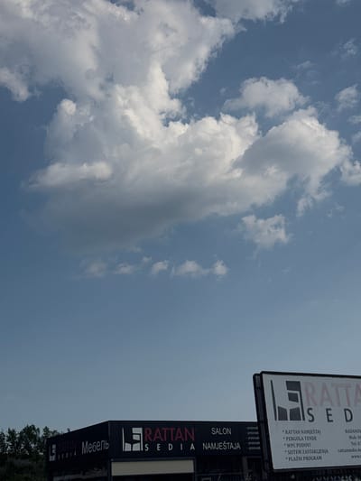 This media file shows a sky with clouds and two signs. The signs are for "RATTAN SEDIA," which appears to be a furniture and outdoor living company. One sign is large and white with black lettering, featuring the company name and a list of products including "RATTAN NAMJEŠTAJ" (Rattan furniture), "PERGOLA TENDE" (Pergola tents), "WPC PODOVI" (WPC floors), "SISTEM ZASTAKLJENJA" (Glazing systems), and "PLAŽNI PROGRAM" (Beach program). It also includes contact information such as a mobile number and website.

Below the larger sign, there is a smaller, darker sign with the same branding. The word "Мебель" (Furniture in Russian) is also visible on one of the signs. The sky suggests it's daytime, possibly overcast or with dramatic cloud formations. The setting appears to be outdoors, with the signs likely placed along a road or in a commercial area. The context provided indicates the location is Dub, Montenegro. There are no people visible in the image.