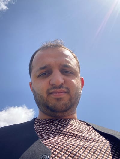 A low-angle, close-up image depicts an adult male subject positioned against a bright blue sky in Nakhonay, Afghanistan. The subject has short, dark, wavy hair and a short, trimmed beard. He wears a patterned shirt featuring a diamond or checkered design in shades of brown and black, beneath a plain black outer garment, possibly a jacket or vest, with one visible black button. The subject maintains direct eye contact with the camera, exhibiting a neutral facial expression. The background consists primarily of a clear blue sky, with a small, diffused white cloud visible in the upper left quadrant. A prominent sun flare is present in the upper right portion of the frame, indicating strong overhead sunlight.
