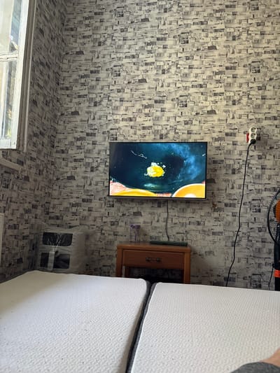 This is a photograph of a room in Tarasovsky, Russia. The room has patterned wallpaper and what appears to be a window on the left side. A flat-screen television is mounted on the wall and is displaying a colorful image. To the right of the television is a wooden cabinet with a brass handle. A portion of a white mattress is visible in the foreground on the right side of the image. The room is lit, suggesting it is daytime. There is no visible text in the image.
