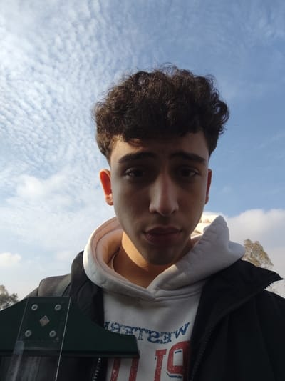 An upward-angled portrait shot depicts a young male subject in Mankat al-Hatab, Syria, looking directly at the camera with a neutral expression. The individual has dark, curly hair and appears to be in his late teens or early twenties. He is dressed in a dark, possibly black, zip-up jacket worn over a light-colored hooded sweatshirt. Partial text "WESTER" in blue and red, along with numerals "19" or "91," is visible on the front of the hoodie. A dark-colored backpack strap is visible over his left shoulder. Sunlight illuminates the right side of his face, casting a shadow on the left. In the lower left foreground, a dark green, rigid object with visible silver fasteners and a square accent partially obscures the subject's torso. The background consists of a clear blue sky densely populated with small, scattered white clouds. Tree foliage is visible in the far background on the right side of the frame.