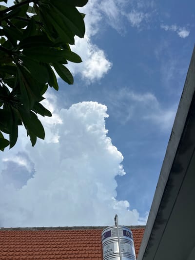 The scene, located in Kerobokan, Indonesia, captures an upward exterior view under a clear daytime sky. The sky is predominantly blue, populated by scattered white cumulus and wispy cirrus clouds. In the upper left quadrant, a cluster of dark green foliage from a tree branch partially obscures the sky. Below the main cloud formation, a section of a residential or commercial building's roof is visible, constructed with reddish-brown terracotta tiles. Positioned on this tiled roof is a large, cylindrical stainless steel water tank, identifiable by "PROFIL TANK" branding and other labels, with a metallic pipe extending vertically from its top. A flat, dark gray architectural element, consistent with a concrete eave or overhang, runs vertically along the right edge of the frame. No individuals are present, and no actions are depicted.