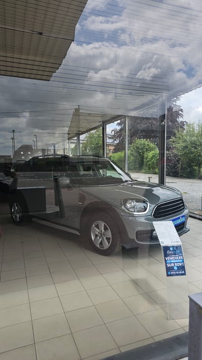 An exterior shot, taken through a window, shows a grey Mini Countryman on display in what appears to be a car dealership in Boussu-lez-Walcourt, Belgium. The car is positioned at an angle, with its driver's side door open. The car's interior is visible, as are its headlights and grille. A sign in front of the car displays the price "€ 14.499,-" and contact information. The car is parked on a tiled floor. Reflections on the window reveal a cloudy sky and the dealership's roof structure. Outside the dealership, trees and buildings can be seen.