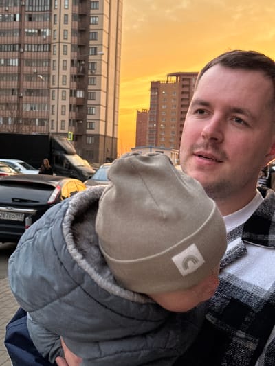 An outdoor scene in the city of Gribovo, Russia, depicts a man holding a baby or young child during golden hour. The man, with short brown hair and light stubble, wears a white t-shirt under a black and white plaid shirt, looking upwards and to his right with a slightly open mouth. The child, held against the man's chest, has its head turned away, revealing a light tan knitted beanie with a white label displaying a grey rainbow icon, and a grey quilted winter jacket with a visible faux fur lining.

The background features several multi-story residential buildings constructed from light brown and beige materials with numerous windows and balconies. The sky is illuminated with vivid orange and yellow hues, indicative of sunset. In the midground, various parked vehicles are visible, including a dark-colored car with a Russian license plate "B267EO 197" (Moscow region), a dark van, and multiple white cars. A woman in a dark coat stands near the parked vehicles. A street lamp is also present. The environment appears to be a residential urban or suburban area.