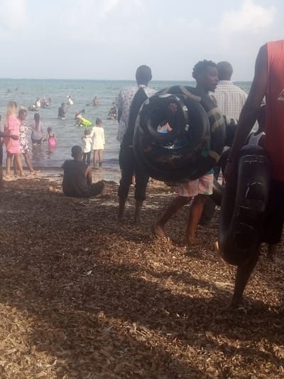This image depicts a crowded beach scene in Shanzu ward, Kenya, under a bright sky. The foreground consists of a shoreline heavily covered with dried brown leaves or seaweed. Several individuals are present:
- On the far right, a person wearing a red sleeveless top carries a dark, inflatable tube under their arm.
- Centrally, a male figure with a dark, wavy hairstyle and patterned shorts carries multiple dark, inflatable tire tubes around his neck and shoulders, walking towards the left.
- To his left, another male figure, seen from behind, wears a light-colored patterned long-sleeve shirt and dark trousers, observing the water.
- In the lower-middle, a male figure in a dark shirt and trousers sits on the water's edge, partially submerged in the shallow water or wet sand, facing the ocean.
- In the midground, several people, including women in dresses (one prominent in pink) and children in light clothing, stand in the shallow water near the shore.
- The background shows numerous people wading, swimming, and floating in the calm, light blue-green ocean. Some individuals in the water are utilizing inflatable devices, including one yellow float visible.
The scene suggests a public recreational area, with people engaged in water activities.