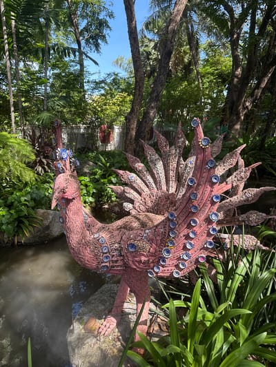 The image depicts a pink, ornate bird-like sculpture adorned with small, blue and white ceramic plates, located in an outdoor garden setting. The sculpture stands beside a body of water, possibly a pond, with lush green foliage surrounding it. Trees are present in the background, with a glimpse of the blue sky visible through the leaves. The location is likely in the city of Samrong Nuea Subdistrict Municipality, Thailand, as stated in the prompt.