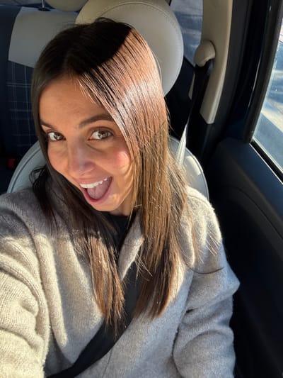 A female subject is photographed in the passenger seat of a vehicle in Antibes, France. The subject has long, straight dark brown hair, brown eyes, and is smiling broadly with an open mouth, exposing her teeth and tongue. She is wearing a light-colored, possibly beige or light grey, knitted cardigan over a dark top, with a black seatbelt secured diagonally across her chest. The vehicle's interior features light-colored seating with a headrest visible behind her. Strong natural light illuminates the scene from the right, casting highlights on her hair and face. A blurred exterior environment is visible through the passenger window, indicating a daytime setting.