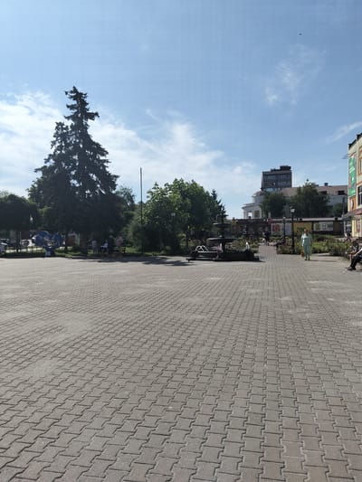 The image depicts a paved plaza in Myrhorod, Ukraine, likely in the city center given the presence of a multi-story building with shops on the ground floor, including one with a banner displaying "200". A prominent feature of the plaza is a multi-tiered fountain with water jets. Several people are visible in the scene: a woman in light-colored clothing walks towards the right, a couple sits on a bench to the left under trees, and other individuals are scattered throughout the plaza, some seated and others standing. Lush green trees, including a large coniferous tree on the left, and flower beds with rose bushes surround the fountain area. The sky is blue with scattered white clouds. The paving stones of the plaza are a neutral gray and extend from the foreground towards the background.