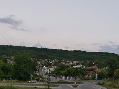 The image depicts a scenic view of a residential area nestled on a green hillside, possibly on the outskirts of Balchik, Bulgaria. The sky is overcast with soft, muted clouds, suggesting it is either early morning or late evening. Power lines are visible stretching across the upper left portion of the frame.
In the foreground, a road intersection with multiple lanes is present. Several traffic signs are strategically placed, including directional arrows and yield signs, indicating a complex junction. A contemporary metal sculpture, abstract in design, stands in the center of the roundabout.
On the left side of the road, a car is visible driving. Further back, a small cluster of buildings, potentially a gas station or a small business, is situated.
The hillside is densely covered with lush green trees and vegetation. Scattered amongst the greenery are numerous houses with red-tiled roofs, characteristic of Mediterranean architecture. The houses vary in size and style, creating a picturesque village-like atmosphere. The overall impression is one of a tranquil and scenic environment, likely a residential neighborhood offering views of nature and the sea, common in the Balchik region.