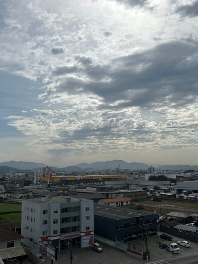 This is a vertical, eye-level aerial shot of a Brazilian city, likely Palhoça, Brazil, with a cloudy sky dominating the upper half of the frame. The clouds are textured, with patches of bright sunlight filtering through, suggesting a partly cloudy day. The lower half of the image shows a dense urban landscape with buildings of varying heights and architectural styles, commercial signage, and parked vehicles.

Several businesses are visible, including "Fort ATACADISTA," "Suvinil," "Osmocolor," and "Construcolor," indicating a commercial district. One building prominently displays "MAIS DO CRISTO," a religious affiliation. The buildings are packed closely together, with some featuring multiple stories and balconies. Parking lots and streets are visible, with several cars, including white pickup trucks and smaller cars, parked. The scene appears to be a busy commercial and residential area.

In the background, distant mountains are visible, their outlines softened by atmospheric haze, suggesting the city is nestled within a valley or near a mountainous region. The lighting and cloud cover indicate it could be late morning or afternoon. The overall impression is a bustling, sunlit urban environment under a dynamic sky.
