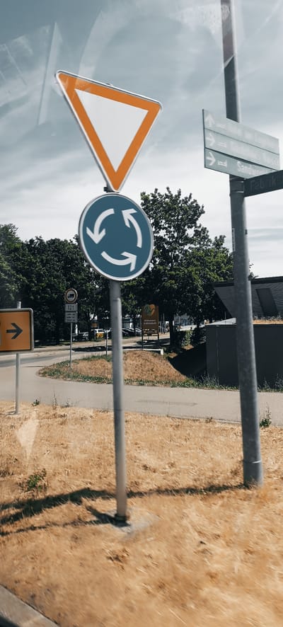 The image shows a collection of road signs on a cloudy day. The main subject is a circular blue sign with three white arrows indicating a roundabout, situated on a grey metal pole. Above this, a large triangular white sign with an orange border, a "Give Way" sign, is also mounted on the same pole. To the right of the roundabout sign, a grey directional sign with white text and arrows indicates "Hotel am Enzpark", "Gästehaus Raitach", and "Sandwerk", as well as a sign for "Fabrikstraße". Further to the left, a yellow square sign with a black arrow pointing to the right is visible. Below the roundabout sign, there is another sign with a truck symbol and "12 t" indicating a weight limit, and some German text below that. In the background, a lush green treeline is visible, with a modern building with a green roof. A small sign with the text "Willkommen in Winterbach" (Welcome to Winterbach) is also present in the background. The ground in the foreground is covered with dry, brown grass, indicating a warm or dry season. The overall scene depicts a road junction in the vicinity of Winterbach, Germany.