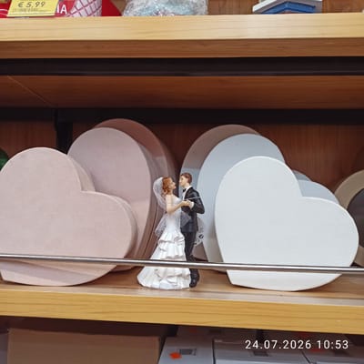 A wedding cake topper featuring a bride and groom is displayed on a shelf in a store in Modugno, Italy. The topper depicts a couple in formal attire, with the bride in a white wedding dress and veil, and the groom in a black tuxedo. They are positioned in front of a collection of heart-shaped gift boxes in various pastel colors. The shelf is made of wood, and behind the boxes, a dark background is visible. A metallic bar runs horizontally across the foreground, partially obscuring the gift boxes. In the upper left corner, a price tag reads "€ 5.99". The bottom right of the image shows a timestamp: "24.07.2026 10:53".