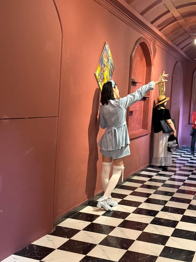 The image shows an interior space with an architectural illusion. A woman in a light blue dress and white stockings is lying on a flat surface with a colorful painting. Another woman in a hat and dark clothing stands nearby. The background features a checkered wall. It is impossible to determine the location in Baqiao, China, based on the image.