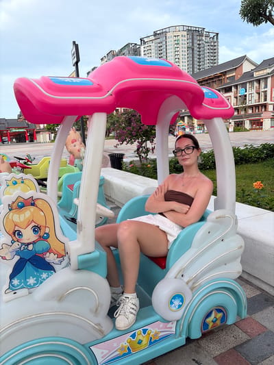 A young woman with dark hair, wearing glasses, a brown strapless top, and white shorts sits in a colorful children's amusement ride shaped like a car with a pink canopy. She has her arms crossed. The ride features a cartoon character, Princess Peach, on the left side. The background shows a street scene with buildings and trees. This scene is located in Nghĩa Trụ Commune, Vietnam.