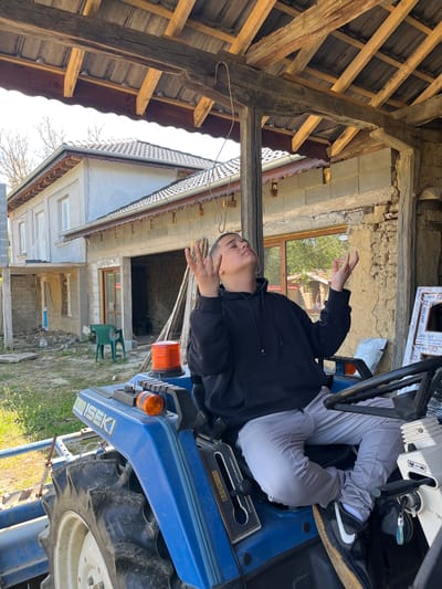 The image captures a person seated on a blue ISEKI tractor, likely in a rural setting. The person is wearing a black hoodie and light grey pants. The background features a partially renovated or under-construction building, with exposed brick and concrete blocks. There is also a wooden roof structure visible, along with a glimpse of vegetation, indicating an outdoor location.
Based on the text "ДОНЕЕ ОРМАТ," "ДОНЕЕ ОРМАТ NOGIH," and "Weiss profil" which may refer to construction materials and the presence of the Cyrillic alphabet, it is plausible that the location could be Rish, Bulgaria, as specified. The construction material manufacturer Weiss Profil operates in Bulgaria. However, without further information or distinct location markers, confirmation is limited.