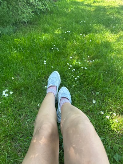 The image shows a person's legs from the knees down, standing on a lawn. The person is wearing white sneakers and pink striped socks. The lawn is green with scattered daisies. The background includes a bush and trees. The location is Velburg, Germany.