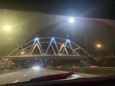 Captured from a vehicle, the image showcases a multi-lane highway at night. A pedestrian bridge with a distinctive blue support structure spans above, adorned with bright lights. Streetlights illuminate the road, where car headlights and taillights create streaks of motion. Graffiti marks the bridge supports. The windshield partially obscures the view.