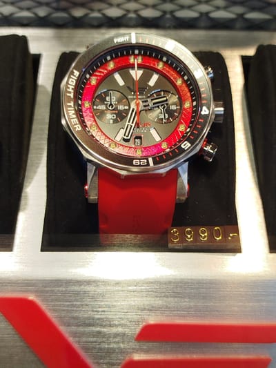 A close-up, eye-level shot captures a dynamic red and silver chronograph watch with a red silicone strap, positioned on a black velvet stand. The watch face displays intricate detailing, including sub-dials, red accents on the bezel and inner ring, and the text "FIGHT TIMER" etched along the outer edge. Below the watch, a wooden price tag with gold-colored numbers reads "3990-". The background is blurred, suggesting an indoor setting, possibly a retail environment. The overall impression is one of a high-end product on display. The scene is forensic and matter-of-fact, devoid of any context regarding its location in Marki, Poland.