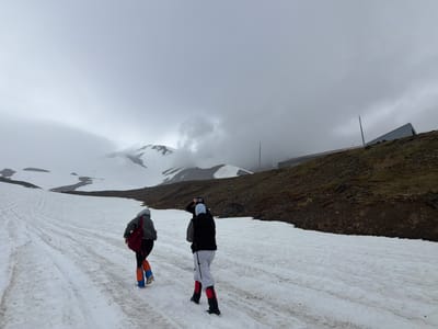 Three individuals are walking up a snowy path towards a building on a hillside. The person in front is wearing a grey hoodie, black pants, and bright orange and blue gaiters. They have a red backpack. The person in the middle is wearing a black vest over a grey hooded sweatshirt, light grey sweatpants, and red and black gaiters. They are holding a walking stick and appear to be adjusting something on their head. The person in the back is wearing a light blue head covering and a dark vest. The ground is covered in snow with tracks from previous footsteps. In the background, there are snow-covered mountains partially obscured by clouds, and a long, modern-looking building with a metal roof situated on a dark, vegetated hillside. The sky is overcast and cloudy, suggesting a cool or cold environment. The overall scene appears to be a hike or exploration in a mountainous, possibly remote, location. There is no visible text in the image.