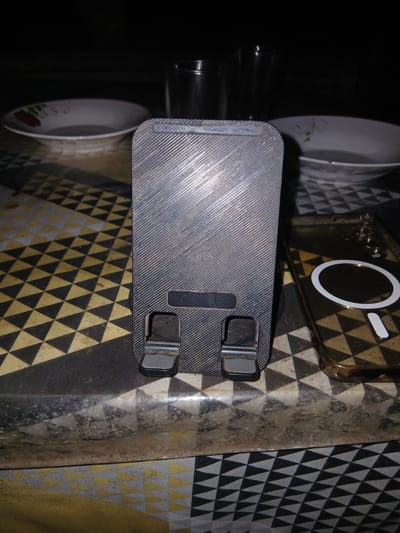 The image depicts a close-up, overhead view of a table surface with a patterned tablecloth. In the foreground, a dark gray, textured phone stand is positioned vertically. To the right of the stand, a transparent phone case with a white circular magnet is visible. In the background, two bowls and two glasses are present, all set on the patterned tablecloth. The lighting is dim, suggesting an indoor environment. There is no discernible urban context or indication of a specific city like Teungkudibandapirak, Indonesia. The scene appears to be a still life arrangement of common household items.