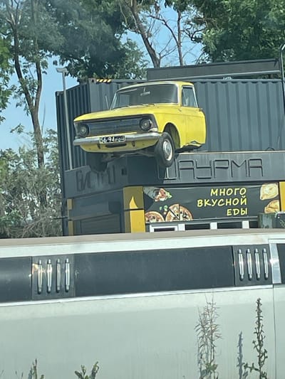 The image shows a yellow car, likely a Moskvich, elevated on top of a large industrial shipping container, which is part of a food establishment. The shipping container is painted dark gray and has the word "ШАУРМА" (Shawarma) in large, white, block letters across its front. Below this, a black banner displays the text "МНОГО ВКУСНОЙ ЕДЫ" (Lots of tasty food) in yellow, stylized lettering, along with images of shawarma, pizza, and some baked goods. The car's license plate is visible, reading "56-94 64". The establishment appears to be located in an outdoor, urban environment with trees and a bright sky. In the foreground, the side of a white vehicle with black trim and chrome accents is visible, suggesting the photo was taken from another vehicle. The location is identified as Kirovsky District, Russia.