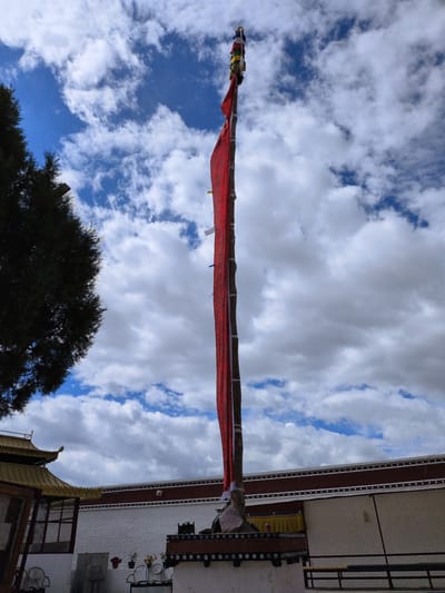 This is a vertical, full shot photograph of a prayer flag pole with a long red flag attached, set against a blue sky with white, puffy clouds. The flag pole is positioned slightly to the left of the center of the image. At the very top of the pole, several smaller, colorful prayer flags are visible. The red flag is long and billows slightly. In the background, a white building with dark red trim and a golden roof can be seen. The building appears to be part of a temple or monastery. A dark green tree with dense foliage is visible on the far left of the image, partially obscuring the sky and the building. The scene is set outdoors, with a bright sky indicating daytime. No people are visible in the frame. The location is Choma, India, as indicated by the presence of prayer flags and traditional Tibetan/Himalayan architecture.