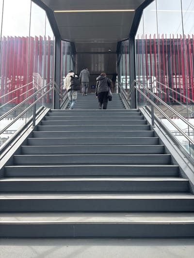 The image depicts an upward view of a modern staircase. The steps are gray and feature vertical bars. A glass and metal structure encloses the staircase, offering views of a nearby building with red horizontal elements. Three people are visible on the staircase, moving upwards. The location is Pétange, Luxembourg.