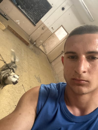 A selfie photograph shows a young man with a shaved head wearing a blue T-shirt in what appears to be a dilapidated building. The man's face is slightly sweaty. In the background, there is a room with peeling paint, a chalkboard, and multiple doors, suggesting a former school or institutional setting. In the foreground, there is a small dog looking up at the camera. The location is suggestive of neglect and possible abandonment. There is no evidence in the photo to indicate that it was taken in Kalchevo, Bulgaria.