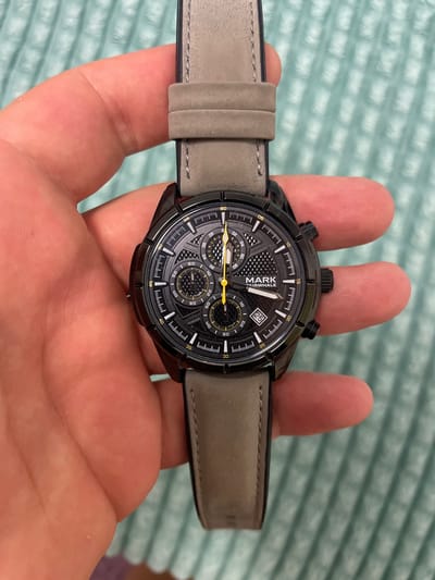 A black analog watch with a grey leather strap is held in a person's hand. The watch face has multiple sub-dials and is black with white markings and yellow accents. The brand name "MARK FAIRWHALE" is visible on the watch face. The background consists of a textured light blue surface. The image does not contain any information that would allow for the determination of its location in Slobozia-Dușca, Moldova.
