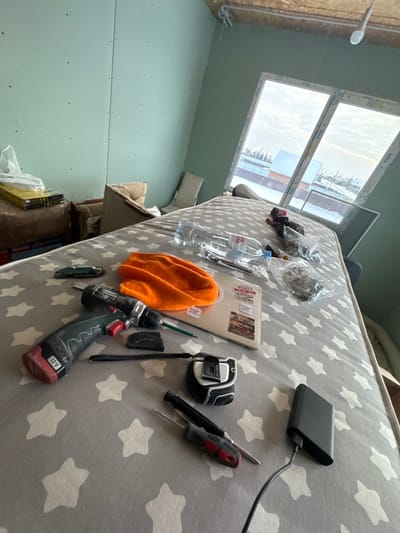 The image depicts an indoor scene within a room under construction or renovation in Novye Chernitsy, Russia. The room features unfinished light green drywall panels on the walls and an exposed wooden beam ceiling with a hanging light fixture.

In the foreground, a light gray mattress with white star patterns serves as a makeshift workspace. Arranged on the mattress are various tools and objects: a black power bank connected via a cable, two screwdrivers (one black/red, one gray/black), a black and white tape measure, a black wrist strap, a used Metabo 12V cordless drill, a pencil, a small black tool part, a closed silver laptop adorned with multiple stickers, an orange microfiber cloth, a clear plastic bottle of Evian water, and two clear plastic bags containing small hardware components.

In the midground, behind the mattress, are stacked cardboard boxes and wrapped items on the left. A disassembled or damaged light-colored armchair is visible, leaning against the wall, with another light-colored high-backed chair further back.

The background is dominated by a large, white-framed, multi-pane window on the right wall, offering a view of an overcast sky and a snow-covered outdoor landscape. Distant buildings, possibly industrial or residential structures, and trees are visible through the window. The overall scene suggests a pause in ongoing renovation or construction work. No individuals are present.
