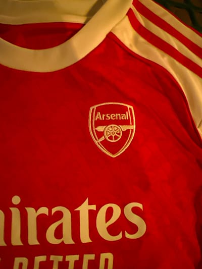 The image shows a close-up of an Arsenal soccer jersey. The jersey is predominantly red with white accents. The Arsenal team logo is visible on the left chest. The Emirates logo is partially visible on the right chest. The collar and stripes on the sleeve are white. The background is out of focus. It's impossible to determine the location in the city of Indian Wells, United States of America from the given image.