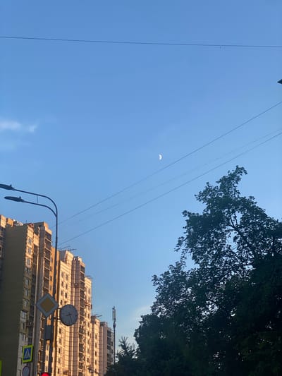 The image depicts a clear blue sky with a crescent moon visible in the upper right quadrant. Several power lines stretch horizontally across the frame. On the left side, a tall building under construction is visible, with its facade partially insulated and windows installed. A street lamp with two fixtures is mounted on a pole in front of the building. Road signs, including a yield sign and pedestrian crossing signs, are present near the base of the pole. A clock is mounted on the same pole, showing approximately 10:10. A cell tower can be seen in the distance behind the building. On the right side of the image, a large, dark green tree dominates the foreground, with its silhouette stark against the sky. The overall scene appears to be an urban or semi-urban environment.