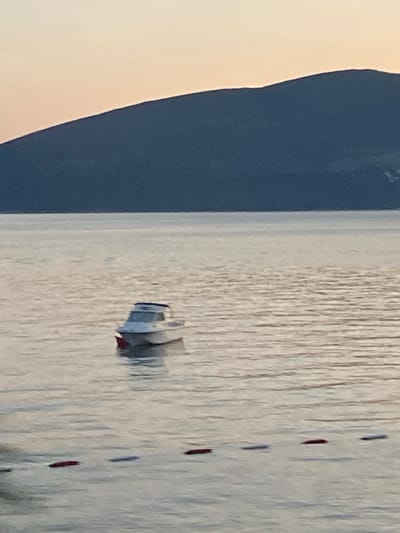 The image depicts a calm body of water with a small white boat with a blue top floating in the middle distance. The water is calm with subtle ripples, reflecting the soft, muted light. In the background, a large, dark blue mountain range dominates the horizon, with a pale, hazy sky above. In the foreground, a line of dark red buoys is partially submerged in the water, marking a boundary. The scene suggests a serene, natural setting, likely at dusk or dawn due to the soft lighting and muted colors, possibly in or near the city of Kumbor, Montenegro, a coastal town known for its scenic beauty and tranquil waters. There are no people visible in the image.