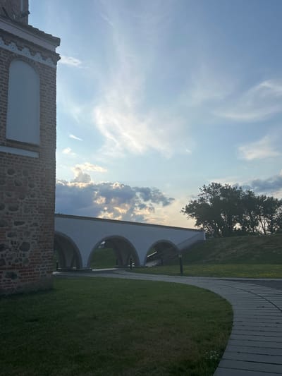 This image captures a scene in Zhukhavichy, Belarus. The foreground features a curved pathway made of grey rectangular stones, winding through a grassy area. To the left, a portion of a brick building with stone accents is visible, its texture and architectural details hinting at historical significance. In the mid-ground, a white arched bridge with multiple openings spans across the landscape. Steps lead up to the right side of the bridge, and a lone street lamp is positioned near the pathway. Behind the bridge, a large, leafy tree stands on a grassy mound. The background is dominated by a blue sky with scattered clouds, some illuminated by sunlight, suggesting it is either sunrise or sunset. No people are present in the image.