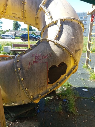 This is a close-up, eye-level, outdoor shot of a large, yellow, plastic tube slide on a playground, located in Gdov, Russia. The slide is weathered and shows signs of wear, with visible graffiti in red marker. A large section of the slide is broken open, revealing the hollow interior and a darker, heart-shaped void. The slide is bolted together with yellow fasteners. In the background, there is a grassy area, a wooden bench, and parked vehicles including a white van and a dark SUV. Behind the vehicles, there are buildings with pitched roofs and a larger, multi-story apartment building. A section of a ladder with yellow rungs and rope is visible on the right side of the frame, attached to a wooden frame. The ground beneath the slide is a dark, rubberized surface, with patches of grass and debris, including some small pieces of trash. The lighting suggests it is daytime with bright sunlight.