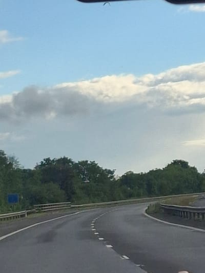 The image shows a view from a moving vehicle on a highway in Darlington, United Kingdom. The road curves to the right, with white dashed lines marking the lanes. Guardrails are visible on both sides of the road, with a blue sign on the left side. Dense green trees line the background, and the sky above is a pale blue with scattered white and gray clouds. The overall scene depicts a typical highway landscape.