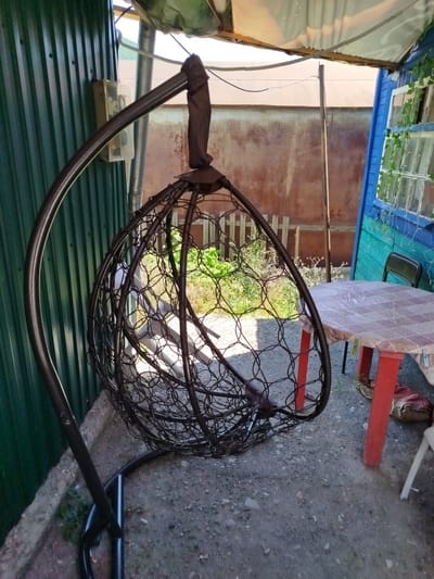 The image shows an outdoor scene in the city of Tuapse, Russia, under a makeshift shelter. In the foreground, a dark metal hanging chair with a circular woven design is suspended from a curved stand. To the right of the chair, a round table with a checkered tablecloth in shades of pink and white stands on red legs. A black chair is partially visible behind the table. The ground is covered with gravel and dirt. In the background, a green corrugated metal fence is on the left, and a blue wooden structure with windows is on the right. Some greenery is visible behind the hanging chair. The scene appears to be an outdoor seating area, possibly in a yard or a patio.