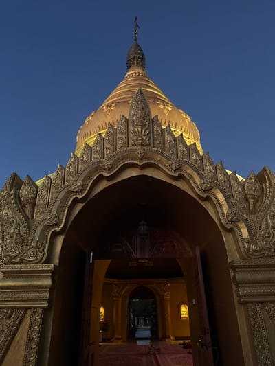 A golden pagoda rises against a clear blue sky. The ornate architecture is prominently displayed, with intricate carvings and layered details. An archway leads into a dimly lit interior, revealing a glimpse of hallways and doorways. Warm light reflects on the walls within the building, creating depth. The composition emphasizes the upward perspective and the grandeur of the structure.