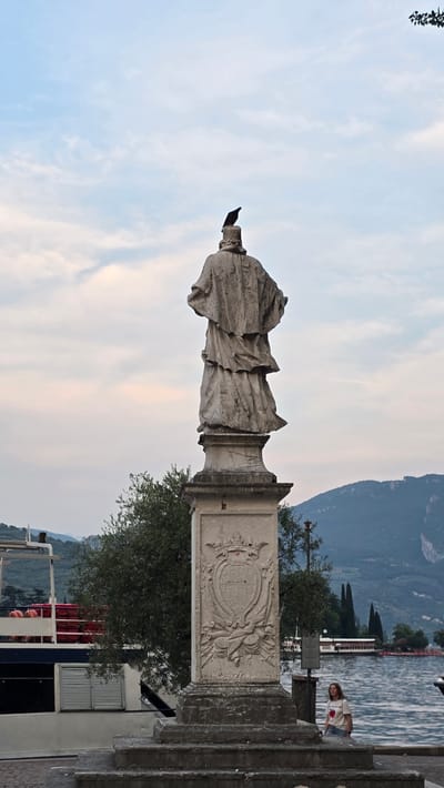 The media file shows a statue of a robed figure standing tall on a pedestal. A bird is perched on the statue's outstretched hand, which is pointing towards the sky. The statue is located outdoors, with a body of water and mountains in the background. The sky is a mix of blue and soft pink, suggesting it might be either sunrise or sunset. The overall scene appears tranquil and serene.

In the background, a body of water, possibly a lake or sea, is visible, with a boat on it. A woman is standing on what looks like a stone walkway, facing the water. The mountains in the distance are covered in lush greenery, and the sky above them is a hazy blue. The statue itself is made of stone and shows signs of age and weathering. The pedestal has an ornate carving on its side, resembling a crest or coat of arms. There is also some greenery around the base of the statue, with trees and bushes adding to the natural setting.

The image's orientation is vertical, suggesting it was taken with a phone or camera held in portrait mode. The lighting conditions indicate it's either early morning or late evening. The overall mood is peaceful and contemplative, with the statue and the natural landscape creating a harmonious scene. The location is suggested to be Riva del Garda, Italy, which is known for its beautiful lakeside scenery and historical sites.
