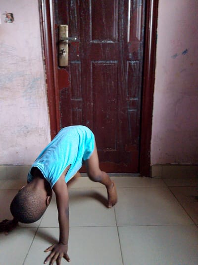 A young child, likely a boy given his short haircut, is captured mid-action in an indoor setting, possibly a home. He is wearing a light blue t-shirt that appears to be a bit large for him, and he is in a downward-facing dog yoga pose or a similar acrobatic position. His head is down, and his hands and feet are on a tiled floor. The floor appears to be made of large, square, light-colored tiles.
Behind the child is a closed, dark wooden door with a brass doorknob and lock. The door has a vertical panel design. To the left of the door, the wall is painted a pale, somewhat textured color, with some scuff marks or fading evident. To the right of the door, the wall is a darker pinkish-purple hue, also with some subtle texture and what might be faint marks.
The lighting suggests it could be daytime, possibly with indoor lighting supplementing any natural light. There are no obvious indications of weather conditions or specific time of day beyond that. The child's pose implies physical activity and playfulness. The scene is simple and domestic, focusing on the child's movement. There is no visible text in the image. The overall impression is a candid shot of a child engaged in energetic play within a typical home environment.