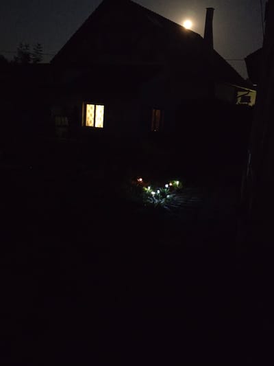 In the city of Kostelec na Hané, Czechia, a nighttime scene is depicted with a silhouetted house under a bright full moon. The house has a dark roof and chimney, with two windows visible. One window on the left glows with a warm yellow light, revealing patterned curtains within. Another window on the right is also lit, though less brightly. In front of the house, a pathway is lined with small, illuminated garden lights that resemble flowers. These lights cast a soft glow on the surrounding grass and the pathway. The overall environment is dark and quiet, suggesting a peaceful rural setting.