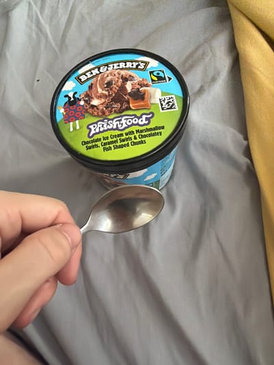 The image shows a pint of Ben & Jerry's Phish Food ice cream sitting on a gray bed sheet, with a silver spoon held in a person's hand next to the container. The ice cream is chocolate flavored with marshmallow swirls, caramel swirls, and chocolate fish-shaped chunks. The location is not determined from the image.