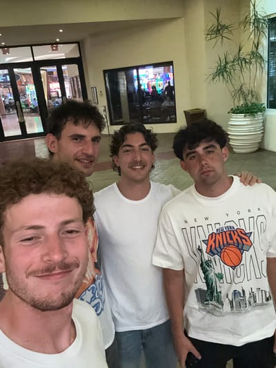 The image shows a group of four young men taking a selfie in what appears to be the lobby of a hotel or resort. The man closest to the camera has curly hair and a light beard. Above him is a man with dark hair and a white t-shirt. Above him is another man with dark curly hair and a white t-shirt. The man at the top is wearing a white New York Knicks T-shirt. The location is likely the Seminole Hard Rock Hotel & Casino Hollywood in Hollywood Seminole Reservation, United States of America, as suggested by the architecture and interior design visible in the background through the glass windows and doorway. A potted plant and a television screen are also visible in the background. They seem to be posing for a casual photo.