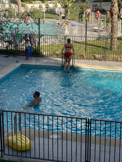 The image depicts an outdoor swimming pool area, likely in a residential or recreational complex. In the foreground, a person is partially submerged in the blue water of the pool, facing away from the camera. To the right, a woman in a red bikini is climbing out of the pool using the steps. The pool is surrounded by a tiled deck and a metal fence. Beyond the fence, several other people are visible, some in the pool and others near the edge. Laundry is hanging on a line in the background. A yellow inflatable object is resting on the deck near the fence in the lower left corner. The environment suggests a warm, sunny day, characteristic of coastal areas like Alicante, Spain, known for its beaches and pools.