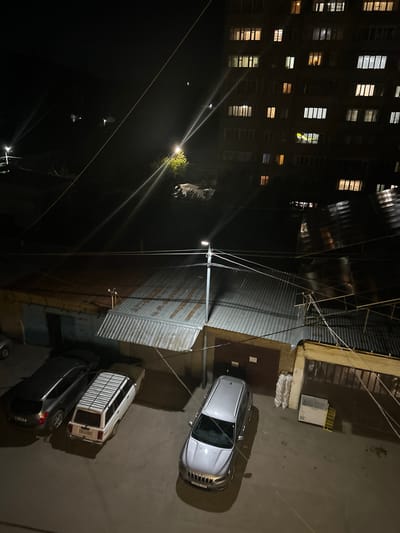The image presents an elevated, static, nighttime view of an urban area in Charentsavan, Armenia. Illumination is provided by various artificial light sources, including streetlights and building windows, which produce prominent lens flares in the upper and middle sections of the frame.
In the upper right background, a multi-story residential building stands dark against the night sky, with numerous windows displaying a mix of warm yellow and cool white interior lights, indicating occupied units. Distant terrain and faint lights are visible in the far left background.
The midground features a series of single-story structures, consistent with garages or storage units. These buildings are characterized by corrugated metal roofs, some with a reddish-brown hue and others with a reflective silver-grey finish. The walls are painted in light, muted colors such as pale blue and beige. A central utility pole supports a complex network of overhead wires that crisscross the frame. Adjacent to a garage on the right, several white sacks, possibly refuse or construction materials, are stacked next to a large white chest freezer or industrial cooler, which in turn is next to a yellow metal rack.
The foreground consists of a paved asphalt surface, serving as a parking area. Three vehicles are parked on this surface. On the far left, a dark grey SUV is visible. Centrally, a white station wagon, equipped with a roof rack, is parked with its rear facing the viewer; its Armenian license plate "34 LA 503" is clearly legible. On the right, a silver SUV is parked, with its front end partially visible and a license plate containing "010 JJJ".
No human figures or active interactions are depicted in the scene. The environment is static, showing residential and utility infrastructure within an urban setting.