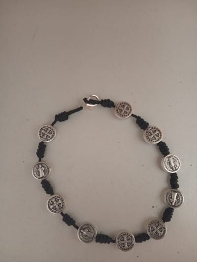 An overhead shot shows a bracelet composed of black cord and silver-colored circular charms. Each charm features a raised design. Some charms display a cross with letters on it, while others depict a figure of a saint. The bracelet is laid out in a circular pattern on a light gray, textured surface. The scene is indoor and the lighting is even. No specific location in Civitanova Marche, Italy can be determined from the image.