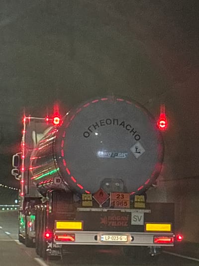 This media file shows a large tanker truck at night. The truck is dark colored with red and yellow reflective markings, and illuminated red and green lights. The tanker itself is cylindrical and gray with red reflective trim. On the side of the tanker, there is text that appears to be in Cyrillic, possibly "ОРТРОНАCHO", and a symbol indicating it carries gas. There are also hazard symbols and numbers like "23" and "1965". On the rear of the truck, a license plate is visible, reading "LP-023-6". The overall scene suggests the truck is parked or moving slowly at night, likely on a road given the reflective markings and lights. The environment appears to be dark, and possibly damp or rainy, contributing to the reflections of the lights. The context provided places the image in Ubisa, Georgia. No people are visible in the image.